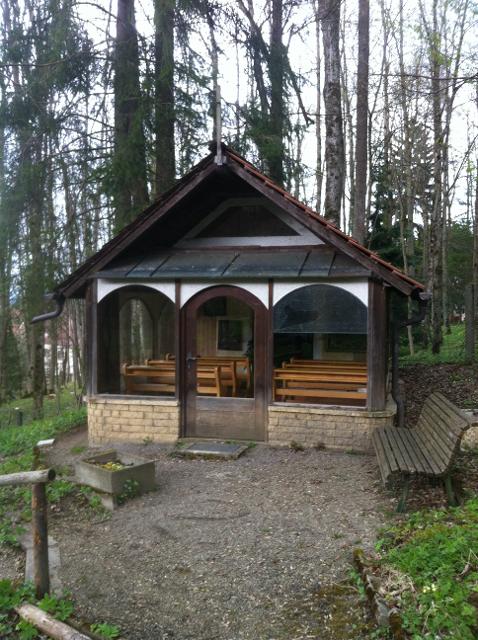 Der Besucher blickt auf eine Kapelle aus dunklem Holz und offener verglaster Front, inmitten eines Waldes.
