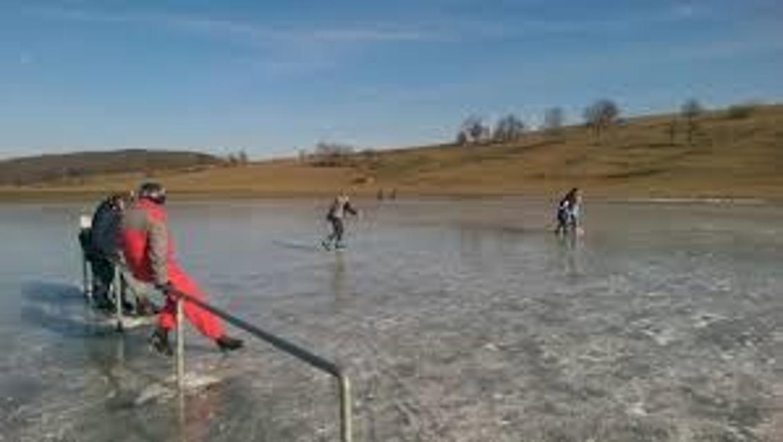 Der Betrachter sieht ein Stück Eisfläche eines Sees und ein Geländer an dem sich Schlittschuhläufer anlehnen.