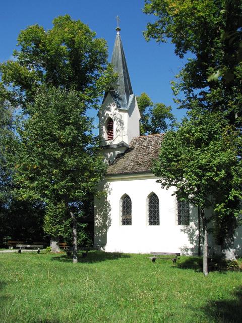 Historische weiße Kapelle auf einer Wiese, eingewachsen von mehreren Laubbäumen.