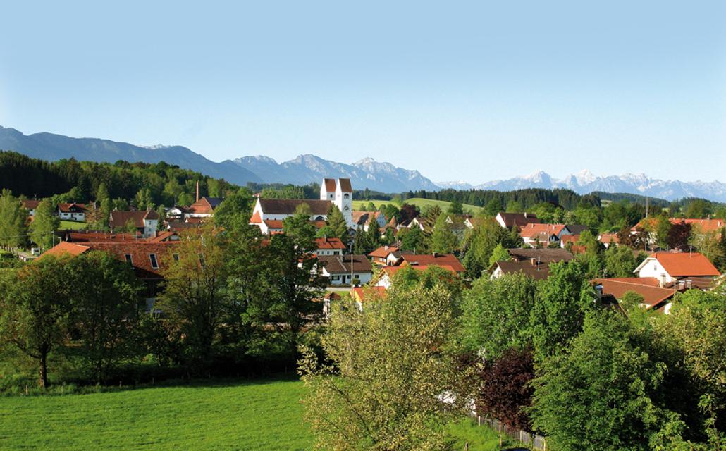 Der Betrachter blickt von einem Aussichtspunkt über eine Wiese hinweg auf das Dorf Steingaden mit seinen typischen Doppelkirchtürmen des Welfenmünsters. Im Hintergrund erhebt sich die Alpenkette.