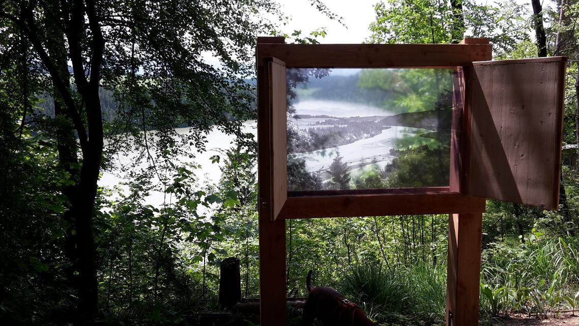 Der Betrachter steht im Wald wo ein Fenster aufgestellt wurde mit einem Glas auf dem der ehemals wilde Lech darauf abgebildet ist, sodass man beim Blick hindurch auf den Lech blickt und die alte Version sieht.