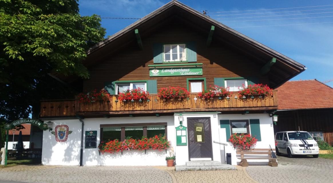 Der Betrachter blickt auf das bayerische Gasthausgebäude mit grünen Fensterläden und Geranien in den Blumenkästen an den Fenstern im ersten Stock.