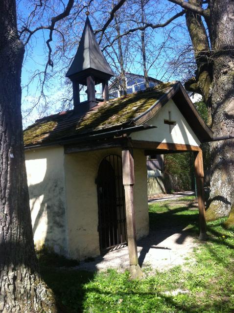 Der Betrachter blickt auf eine Kapelle die zwischen zwei großen Laubbäumen steht.