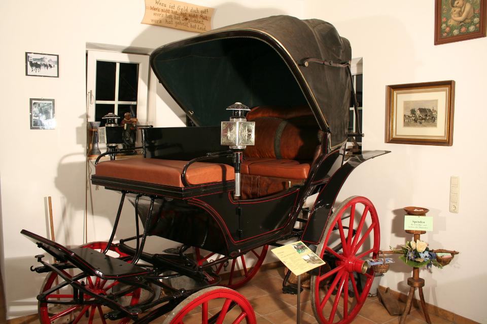 Der Betrachter befindet sich im Museum und blickt auf eine schwarze noble Kutsche mit Überdachung und roten Holzrädern.