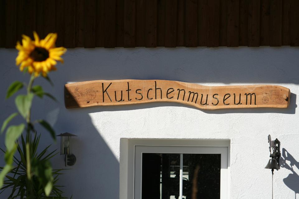 Der Betrachter steht vor dem Eingang des Museums und blickt auf das Holzschild über dem Eingang mit dem Schriftzug "Kutschenmuseum".