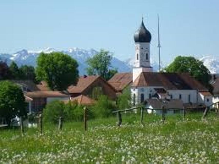 Der Betrachter blickt über eine Frühlingsblumenwiese hinweg auf ein Dorf mit Kirche und Maibaum. Am Horizont erhebt sich die Alpenkette.
