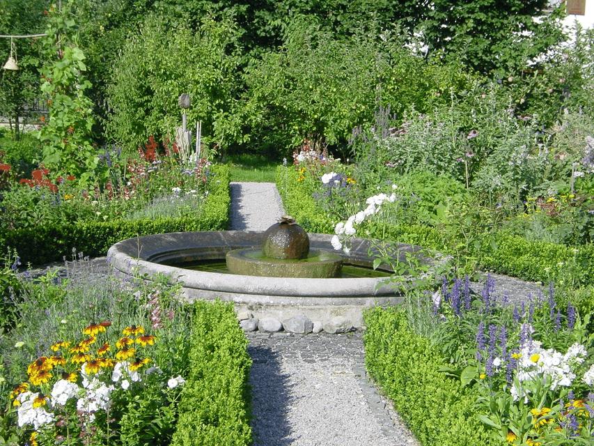 Der Betrachter befindet sich in einem in quadratur angelegten Kräutergarten in welchem sich mittig ein runder, steinerner Brunnen befindet.