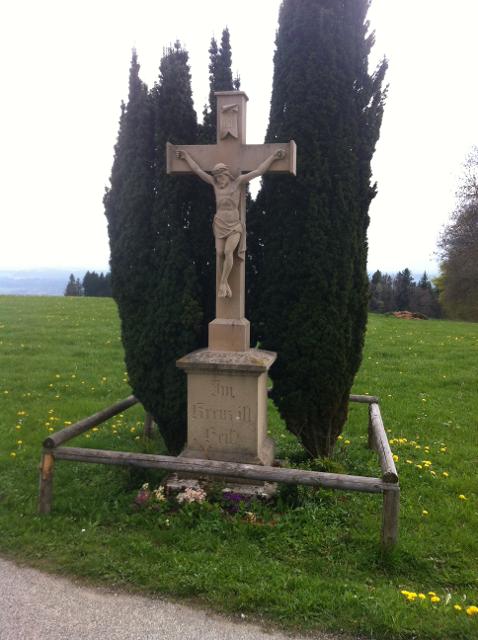 Ein kunstvolles Feldkreuz aus Stein steht zwischen zwei Eibensträuchern in einer weitläufigen Wiese.