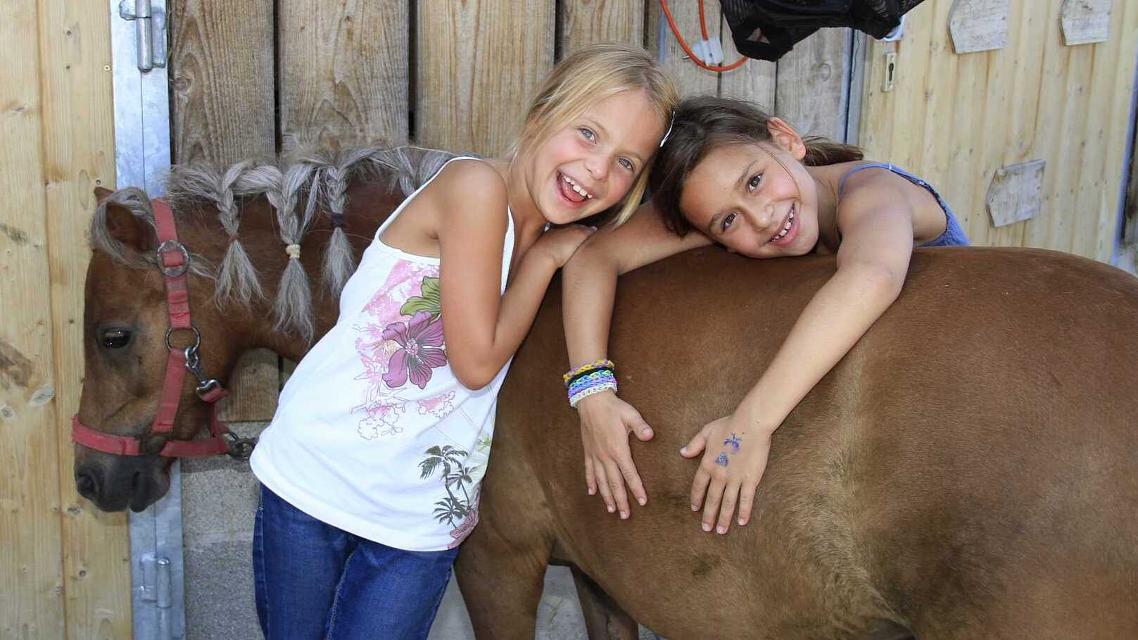 Zwei Mädchen lehnen sich lächelnd an ein Pony bzw. über den Ponyrücken.