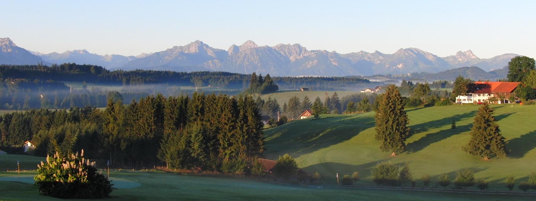 Der Betrachter blickt auf den Golfplatz und das Alpenpanorama am Horizont.