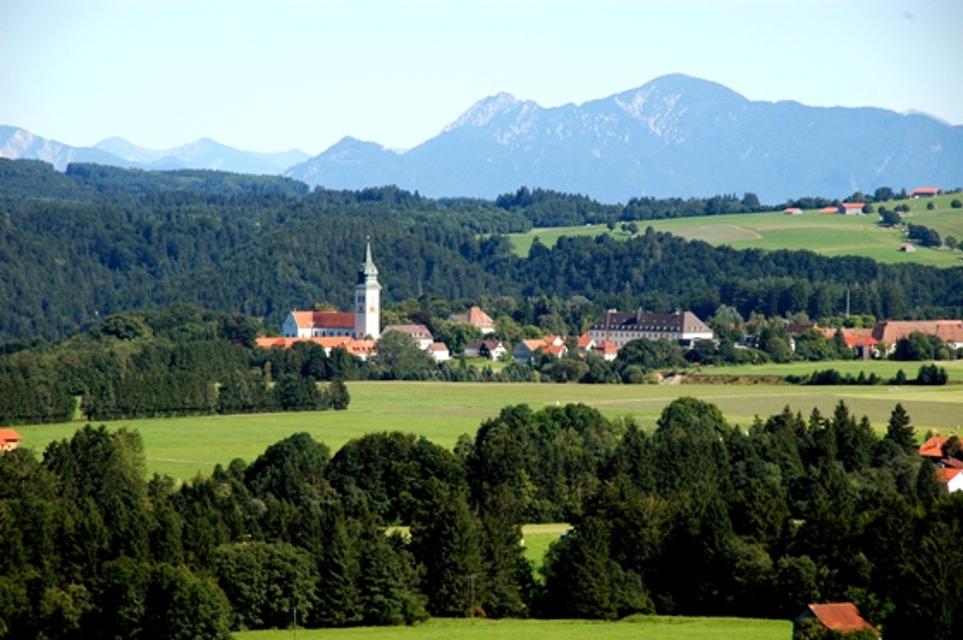 Von der Ferne blickt der Betrachter über die Wiesen und Wälder auf das ehemalige Klosterdorf Rottenbuch mit seinem 67m hohen Kirchturm. Im Hintergrund erhebt sich die majestätische Alpenkette.