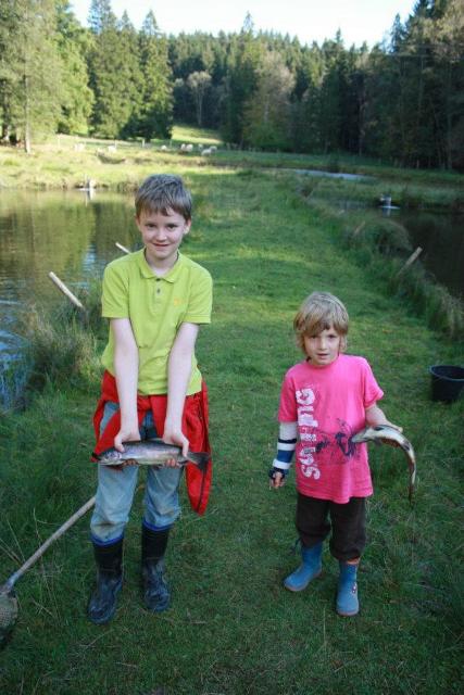 Zwei Kinder halten am Fischteich stehend ihre gefangenen Forellen in den Händen.