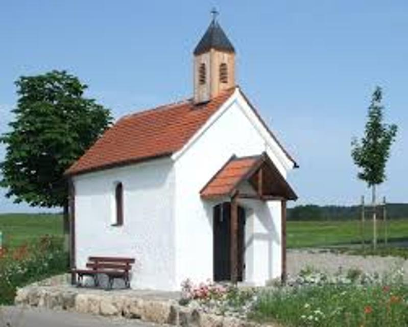 Der Betrachter blickt auf eine weiße Kapelle an deren Außenwand eine Parkbank zum Ausruhen aufgestellt wurde. Die Kapelle steht auf einem Feld.