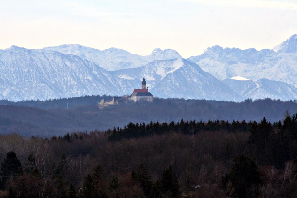 Von der Ferne ist das Kloster Andechs inmitten von Wäldern zu sehen. Am Horizont das grandiose Alpenpanorama.