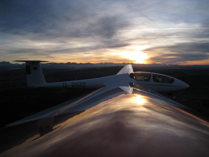Der Betrachter blickt von einem Tragflächenende direkt seitlich auf das Segelflugzeug. Am Horizont geht die Sonne unter.