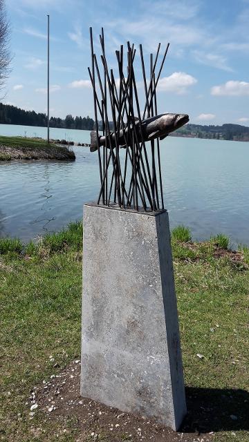 Der Betrachter steht direkt am Lechstausee und blickt direkt vor sich auf ein Kunstobjekt, das einen Fisch aus Metall zeigt, der durch Metallstangen quasi schwimmt.