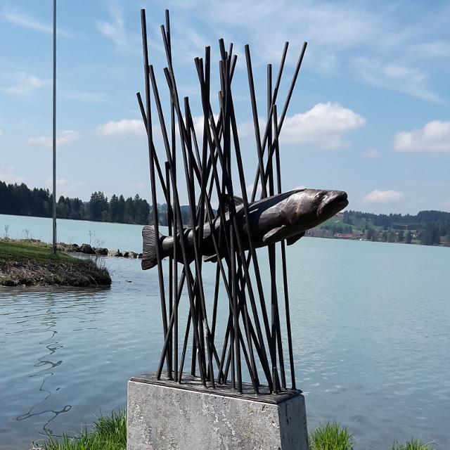 Der Betrachter steht direkt am Lechstausee und blickt direkt vor sich auf ein Kunstobjekt, das einen Fisch aus Metall zeigt, der durch Metallstangen quasi schwimmt.