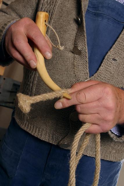 Der Betrachter sieht bei einem alten Handwerk zu. Dabei hat ein Mann ein Seil und eine Art Horn in der Hand.
