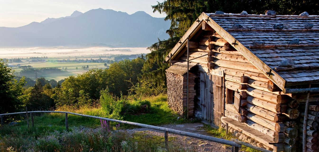 Der Betrachter blickt auf ein historisches Bauernhaus. Im Hintergrund die Alpenvorlandschaft und Berge.