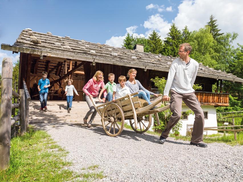 Der Betrachter sieht eine Familie die mit einem alten Holzschubkarren die Kinder transportieren. Im Hintergrund ein historisches Bauernhaus.