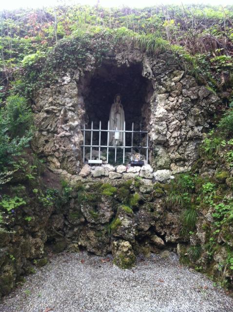 Der Betrachter blickt auf eine steinerne Grotte die sich in einem Berg befindet. Hinter einem kleinen Gitter steht die betende Madonna. Die Steine sind sehr bemoost und mit Efeu teilweise eingewachsen.