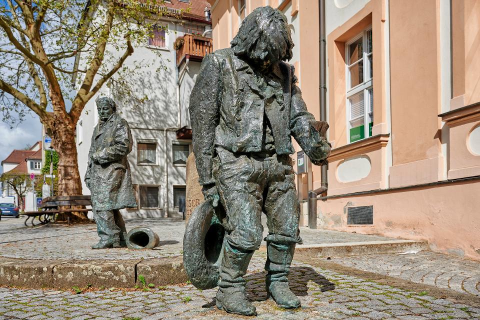 Friedrich Schelle griff eine Idee des Ansbacher Bildhauers Waldemar Fritsch auf und gestaltete die Bronze-Figuren. Dargestellt ist Kaspar Hauser zweimal: die gebeugte Gestalt zeigt ihn, wie er am 26. Mai 1828 in Nürnberg aufgetaucht sein könnte...