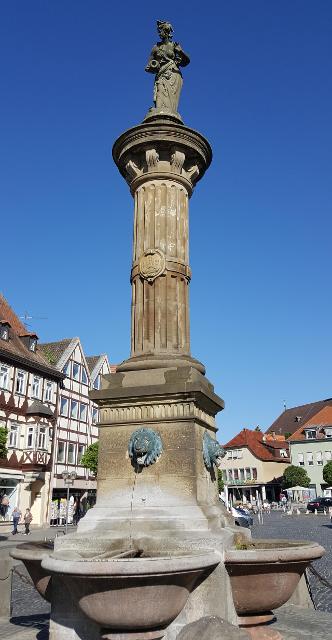 Im Zentrum des historischen Marktplatzes, dem Herzstück der Bad Neustädter Altstadt, steht ein alter Brunnen, der seit 1881 mit einer Figur aus der Barockzeit gekrönt ist.
Die Dame, die da milde lächelnd auf das bunte Markttreiben herabblickt, ste...